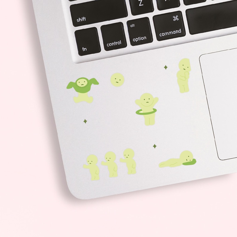 Puede incluir: Un conjunto de pegatinas verde claro con varias figuras de dibujos animados y peque&ntilde;as formas de diamante. Las pegatinas est&aacute;n colocadas sobre una superficie blanca de ordenador port&aacute;til, cerca del teclado, con un fondo rosa claro.