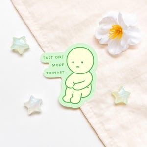 May include: A sticker featuring a seated, light yellow cartoon figure with a sad expression, outlined in green. Text on the sticker reads "JUST ONE MORE TRINKET." The sticker is surrounded by three iridescent star-shaped decorations and a white flower.