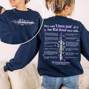 May include: Navy blue sweatshirt featuring a floral and sword design with text. The front has a floral and sword design. The back has text that reads "They said 'I love you' but Kai Azer once said..." and a list of quotes. The sweatshirt is part of "The Powerless Trilogy" by Lauren Roberts.