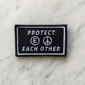 May include: Black embroidered patch with white text that reads "PROTECT EACH OTHER" with a peace sign and the letter "E" in a circle.