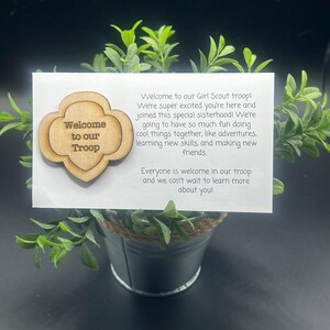 May include: A wooden Girl Scout troop welcome sign with the text "Welcome to our Troop" on a white background. The sign is in the shape of a Girl Scout cookie.