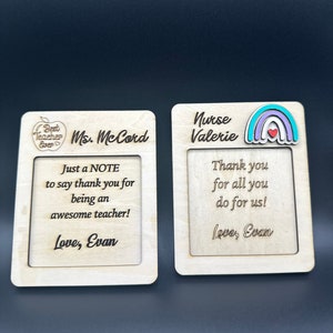 May include: Two wooden cards with engraved messages. The first card says "Best Teacher Ever" and "Ms. McCord" with a red apple graphic. The second card says "Nurse Valerie" with a rainbow graphic. Both cards have the message "Love, Evan".