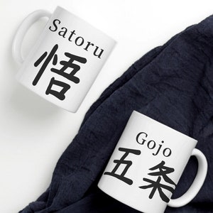 May include: Two white ceramic mugs with black lettering. The mug on the left says "Satoru" in English and "悟" in Japanese. The mug on the right says "Gojo" in English and "五条" in Japanese. The mugs are on a white surface with a dark blue cloth in the background.