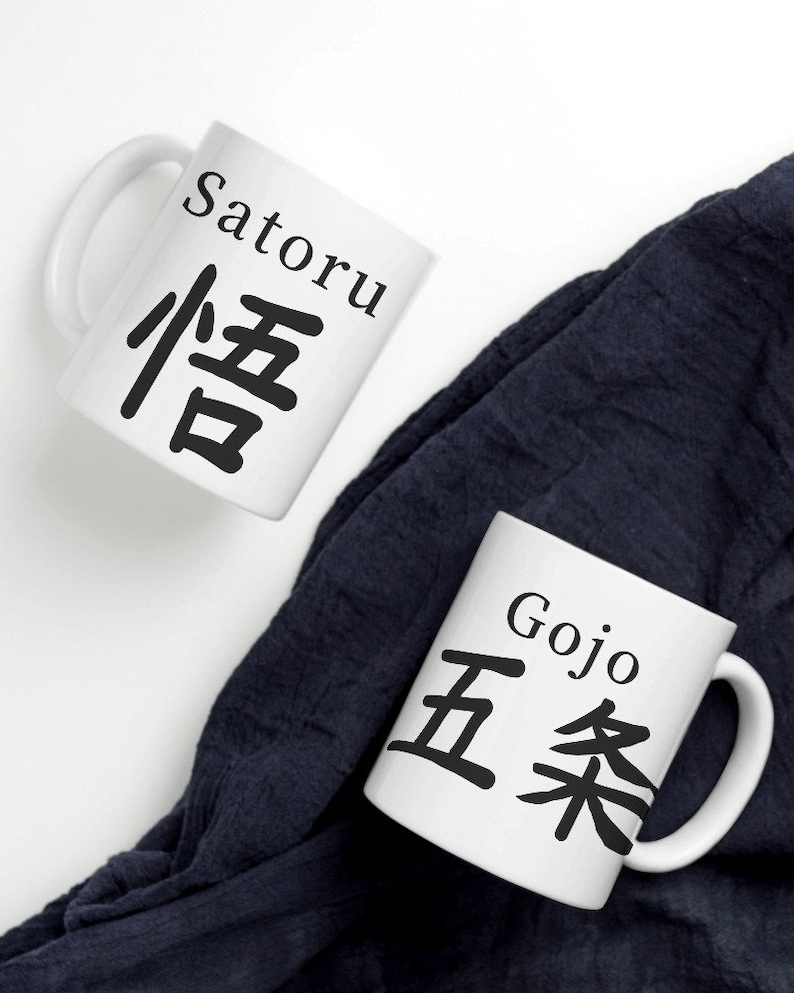 May include: Two white ceramic mugs with black lettering. The mug on the left says "Satoru" in English and "悟" in Japanese. The mug on the right says "Gojo" in English and "五条" in Japanese. The mugs are on a white surface with a dark blue cloth in the background.