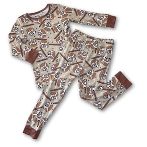 May include: Tan pajama set with a repeating pattern of brown and white cartoon-style dog faces and brown sticks. The long-sleeved top and pants have brown cuffs and trim. The pajamas are made of a soft material.