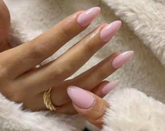 May include: Close-up of a hand with long, almond-shaped, light pink nails. A gold ring is visible on the ring finger. The hand rests on a fluffy, white, faux fur surface.