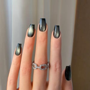 May include: A hand with long, square-shaped nails painted in a dark, iridescent color. The nails have a glossy finish, reflecting light with hints of gold and green. A silver ring with a linked design is worn on the ring finger.