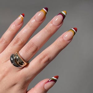Op de afbeelding: Amandelvormige vingernagels met een meerkleurig French tip-ontwerp met strepen in bordeauxrood, wit, mosterdgeel en olijfgroen. Een dikke, gepolijste ring met afwisselende banden van goud- en zilverkleurig metaal zit om de ringvinger.