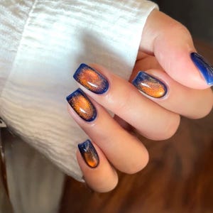 Könnte beinhalten: Nahaufnahme einer Hand mit gepflegten Nägeln. Die Nägel sind mit einer dunkelblauen Basis und einem schimmernden orange-goldenen Cat-Eye-Effekt lackiert. Die Nägel sind quadratisch geformt.