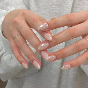 May include: Close-up of hands with a light pink, pearlescent nail polish. The nails are shaped into a long oval and have a glossy finish.