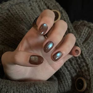 May include: A hand with oval-shaped nails painted in a brown shade with an iridescent shimmer. The nails reflect blue and green hues. The hand rests on a grey knitted jumper.
