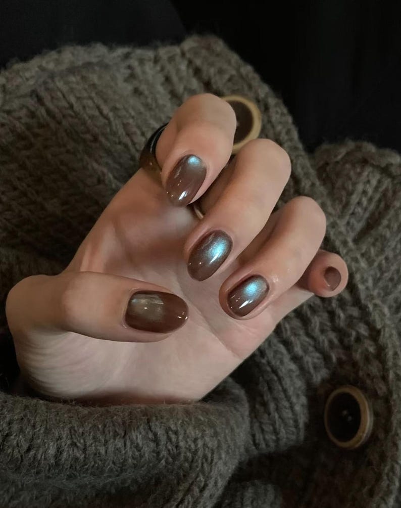 May include: A hand with oval-shaped nails painted in a brown shade with an iridescent shimmer. The nails reflect blue and green hues. The hand rests on a grey knitted jumper.