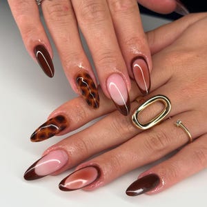 Può includere: Primo piano di una mano con unghie lunghe e a mandorla dipinte con una varietà di disegni. Alcune unghie sono dipinte con un motivo a tartaruga marrone e nero, mentre altre sono dipinte con un effetto ombré marrone e rosa. Un'unghia è dipinta con un colore marrone solido.