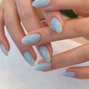 Könnte beinhalten: Nahaufnahme von Händen mit oval geformten, hellblauen Nägeln. Die Nägel haben ein glänzendes Finish und einen subtilen Farbverlauf, der von einem tieferen Blau am Ansatz zu einem helleren Farbton an den Spitzen übergeht. Die Nägel sind gut gepflegt.