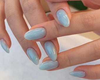 Könnte beinhalten: Nahaufnahme von Händen mit oval geformten, hellblauen Nägeln. Die Nägel haben ein glänzendes Finish und einen subtilen Farbverlauf, der von einem tieferen Blau am Ansatz zu einem helleren Farbton an den Spitzen übergeht. Die Nägel sind gut gepflegt.