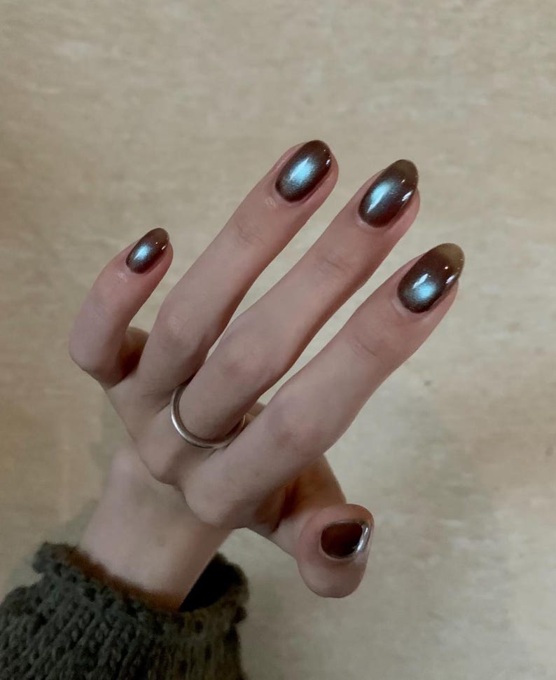 May include: A hand displaying oval-shaped nails painted in a dark brown shade with a blue iridescent effect. The nails have a glossy finish and reflect light. A silver ring adorns the ring finger. The background is a light beige colour.