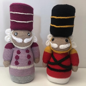 May include: Two crocheted nutcracker dolls, one in purple and gray with a burgundy hat and the other in red and white with a black hat.