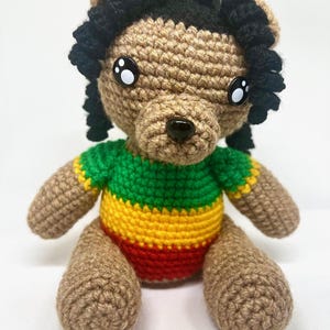 May include: A crocheted teddy bear with a brown body, black dreadlocks, and large eyes. The bear is dressed in a green, yellow, and red striped top. The handmade toy is designed to be soft and cuddly, a charming gift.