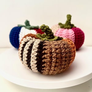 Peut inclure: Trois citrouilles crochetées dans les tons de marron, beige et orange, avec des tiges vertes. Deux citrouilles supplémentaires sont visibles en arrière-plan, une bleue et blanche, l'autre rose et bordeaux. Les citrouilles sont exposées sur une surface blanche.