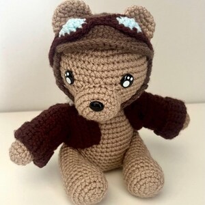 Może przedstawiać: Pluszowy miś w kolorze brązowym, wykonany na szydełku, w brązowej czapce lotniczej z niebieskimi akcentami i brązowej kurtce.