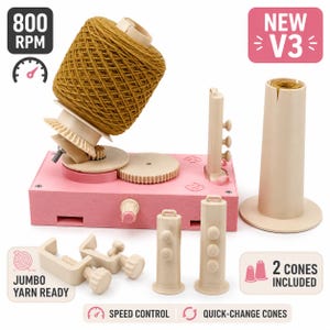 Op de afbeelding: Een roze en beige elektrische garenwinder met een mosterdgele garenkegel, voorzien van de teksten 800 RPM, NEW V3, JUMBO YARN READY, 2 CONES INCLUDED, SPEED CONTROL en QUICK-CHANGE CONES. Inclusief twee beige klemmen en twee beige kegelhouders.