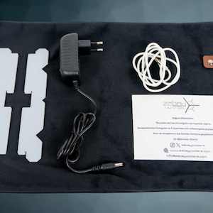 May include: A black velvet pouch with a brown leather tag that says "zelbou" contains a white light panel, a black power adapter, a white cable, and a white instruction sheet with the text "zelbou" and contact information.