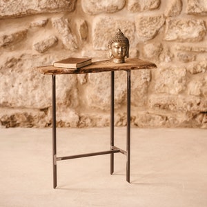 Może przedstawiać: Mały drewniany stolik z blatem o żywym brzegu i czarnymi metalowymi nogami. Na blacie leży książka i złota statuetka Buddy.