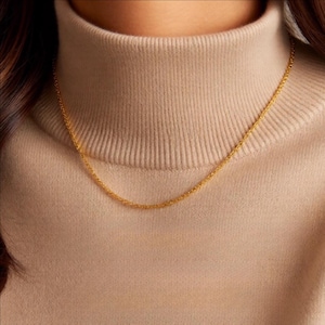 Op de afbeelding: Een fijne, goudkleurige ketting. De ketting wordt gedragen tegen een beige coltrui. De ketting heeft een delicate, getextureerde uitstraling en de algehele esthetiek is eenvoudig en elegant.