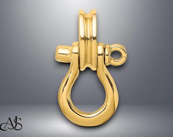 Pode incluir: Um pingente dourado em forma de grilhão. O pingente tem um corpo em forma de U com uma parte superior arredondada e uma argola para uma corrente. O metal tem um acabamento polido, refletindo a luz. O fundo é um gradiente de cinza.
