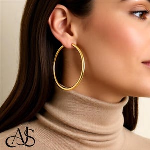 May include: A close-up of a person wearing a large, gold-colored hoop earring. The earring is circular and has a smooth, polished surface. The person is wearing a beige turtleneck sweater. The image has a warm, soft lighting.
