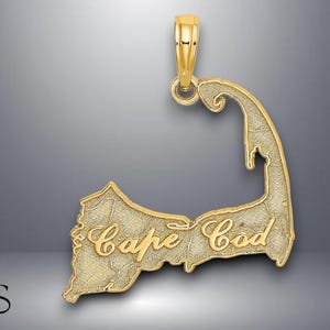 May include: A gold-colored pendant in the shape of Cape Cod, with the words "Cape Cod" in a cursive font. The pendant has a textured surface and a loop at the top for a chain.