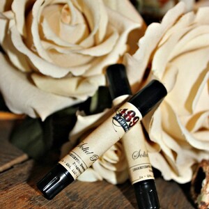 May include: Two small black bottles with labels that say "Solid Perfume" and "1889 Apothecary" are sitting on a wooden surface. The bottles are surrounded by white roses.