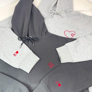 Op de afbeelding: Meerdere grijze en donkergrijze hoodies met rode hart- en kaartsymboolborduurwerk. Eén hoodie heeft een hartpuzzelontwerp. De mouwen tonen de letters K en J met een hart. De hoodies zijn gemaakt van zacht materiaal.