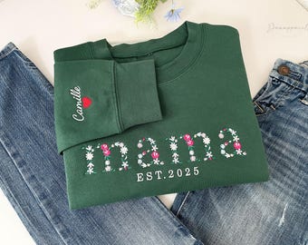 May include: A dark green sweatshirt with the word "mama" embroidered in white with pink and white floral accents. The text "EST. 2025" is below the word "mama". The sleeve cuff has the name "Camille" and a red heart.