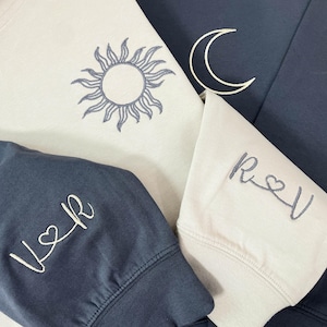Puede incluir: Primer plano de varias sudaderas en tonos azules y crema. Una sudadera presenta un diseño de sol, otra una luna creciente y otras tienen iniciales bordadas. Es probable que las sudaderas formen parte de un conjunto a juego.