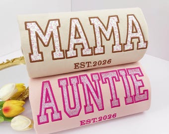 May include: Two decorative pillows, one beige and one pink. The beige pillow has the word "MAMA" embroidered in brown with a floral pattern, and "EST.2026". The pink pillow says "AUNTIE" in glittery pink, and "EST.2026".