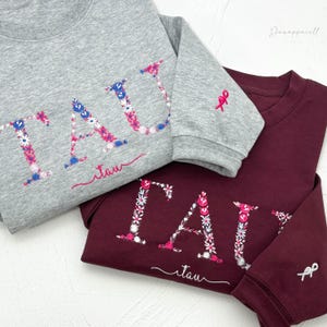 Puede incluir: Dos sudaderas con el nombre "TAY" bordado en flores de colores. Las sudaderas son grises y burdeos.