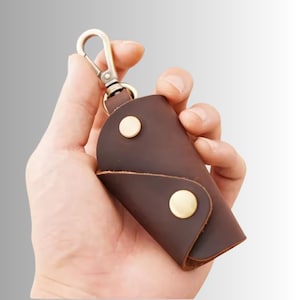 May include: A dark brown leather key case featuring a smooth matte finish, secured with two round brass-coloured metal snap buttons. The case includes a matching brass-coloured metal swivel lobster clasp attached to the top for easy key attachment.