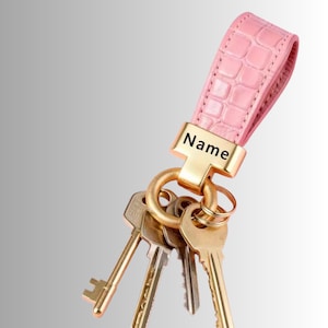 May include: A light pink keyring with a textured crocodile skin pattern and a gold-toned metal clasp. The clasp features the word Name printed in black. A gold-toned metal ring is attached to the clasp, holding four assorted metal keys.