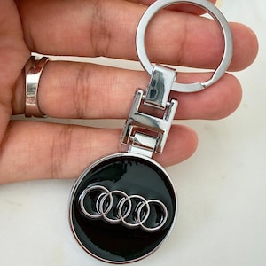 Könnte beinhalten: Ein silberfarbener Metallschlüsselanhänger mit einem runden, schwarz emaillierten Anhänger, der das silberne Audi-Logo mit vier Ringen zeigt. Der Anhänger ist an einem rechteckigen Metallglied befestigt, das mit einem Standard-Schlüsselring verbunden ist.