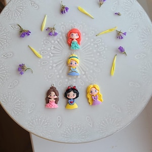 May include: Six princess-themed charms in various colours are displayed on a white, patterned surface, with scattered purple flowers and yellow petals. Each charm features a different princess design.