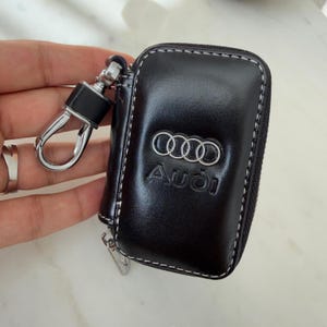 Puede incluir: Un estuche para llaves de cuero negro con el logotipo plateado de Audi y la palabra Audi grabada en el frente. El estuche rectangular tiene costuras blancas en contraste a lo largo de los bordes e incluye un clip giratorio de metal plateado.