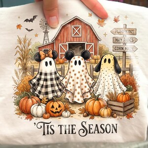 以下が含まれることがあります： 白いTシャツに、納屋、カボチャ、そして「PUMPKINS」「HAY RIDES」「CORN MAZE」と書かれた看板の前で3体の幽霊が描かれたグラフィック。シーンの下には、暗い筆記体フォントで「TIS THE SEASON」というテキストがプリントされています。