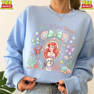 May include: Light blue sweatshirt featuring a colorful graphic of Ariel from The Little Mermaid, surrounded by seashells, bows, and the text "Part Of Your World." A dog and a crab are also included in the design. The sweatshirt has long sleeves.