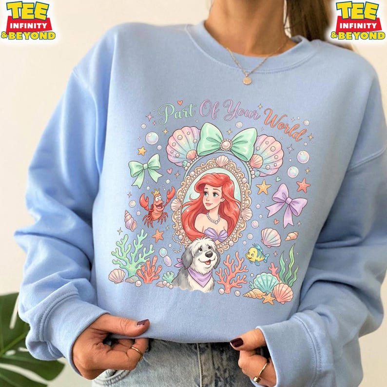 May include: Light blue sweatshirt featuring a colorful graphic of Ariel from The Little Mermaid, surrounded by seashells, bows, and the text "Part Of Your World." A dog and a crab are also included in the design. The sweatshirt has long sleeves.