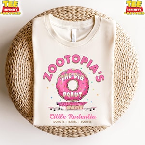 Puede incluir: Camiseta color crema con un dise&ntilde;o de donut rosa y el texto "ZOOTOPIA'S THE BIG DONUT". Debajo, el texto dice "Cittle Rodentia" y "DONUTS - BAGEL - SCOFFEE". La camiseta tiene cuello redondo y mangas cortas.