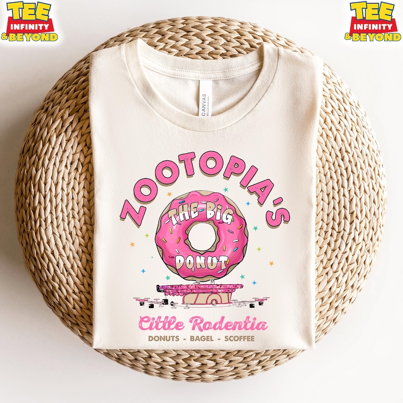 Puede incluir: Camiseta color crema con un dise&ntilde;o de donut rosa y el texto "ZOOTOPIA'S THE BIG DONUT". Debajo, el texto dice "Cittle Rodentia" y "DONUTS - BAGEL - SCOFFEE". La camiseta tiene cuello redondo y mangas cortas.