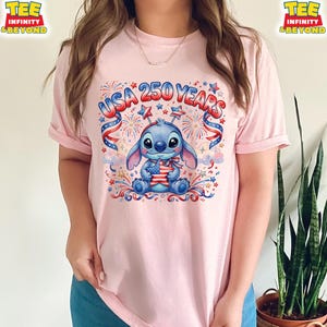 May include: A light pink t-shirt featuring a blue cartoon character holding an American flag. The shirt has the text "USA 250 YEARS" in red and blue, with fireworks and star accents. The shirt is a crew neck style.