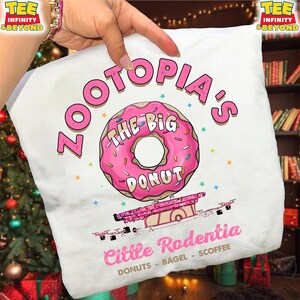Puede incluir: Camiseta blanca con un dise&ntilde;o de donut rosa, chispas y el texto "ZOOTOPIA'S THE BIG DONUT". Debajo, un carrito de comida rosa con "Little Rodentia" y el texto "DONUTS - BAGEL - SCOFFEE".