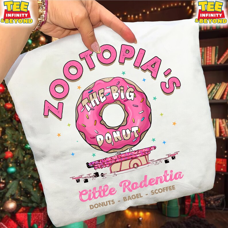 Puede incluir: Camiseta blanca con un dise&ntilde;o de donut rosa, chispas y el texto "ZOOTOPIA'S THE BIG DONUT". Debajo, un carrito de comida rosa con "Little Rodentia" y el texto "DONUTS - BAGEL - SCOFFEE".
